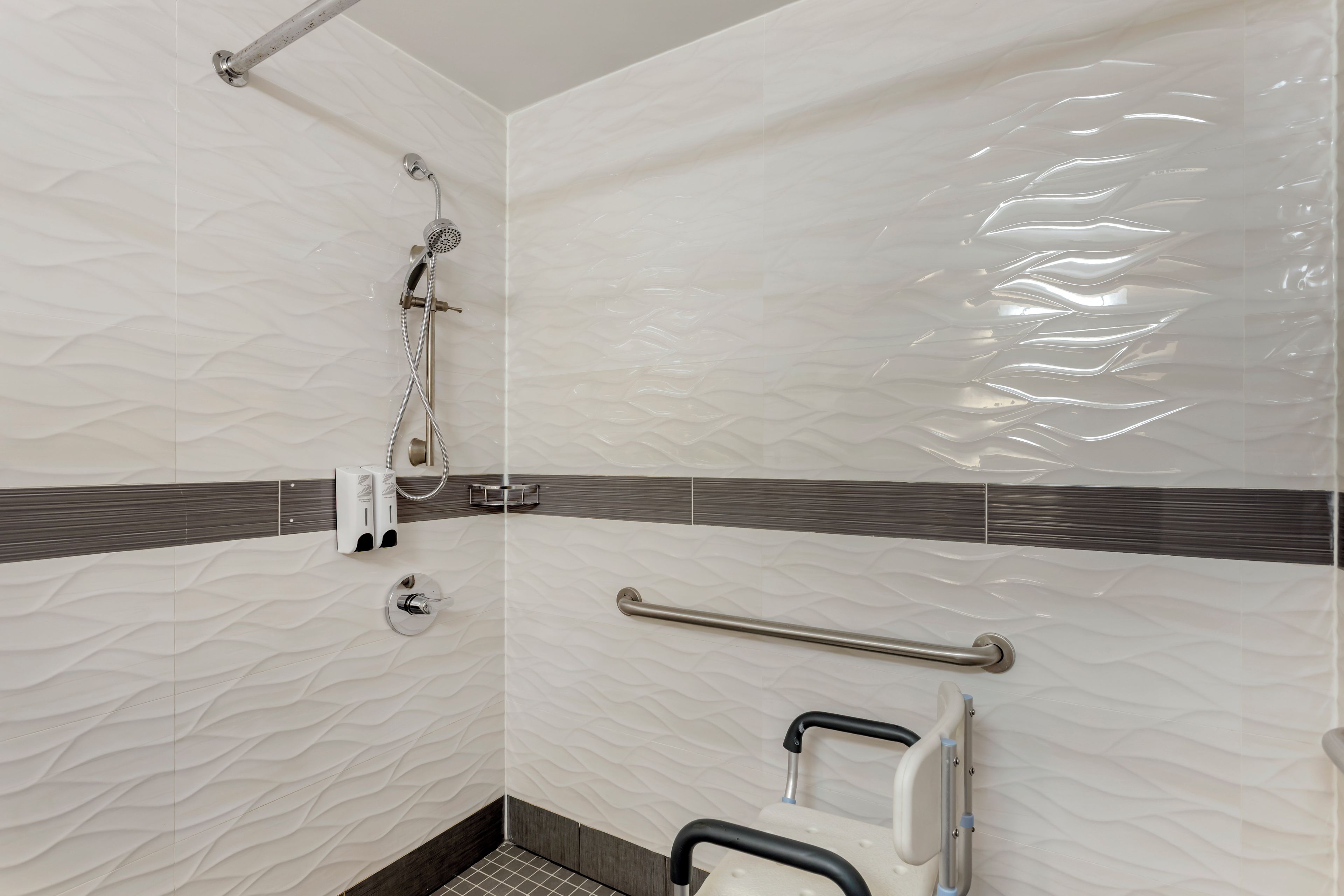 A bathroom with a textured wall, a shower with a handheld showerhead, a grab bar, and a soap dispenser.