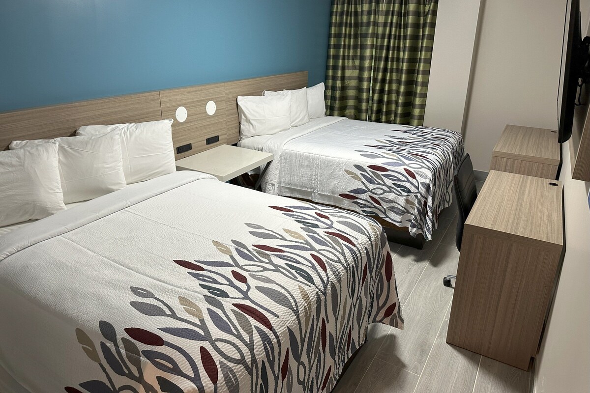 Et hotelværelse med to senge, et lille bord, et træskab og et fjernsyn.