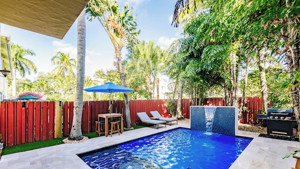 Une piscine avec une terrasse carrelée, une table en bois, deux chaises longues, un parasol bleu et une clôture rouge.