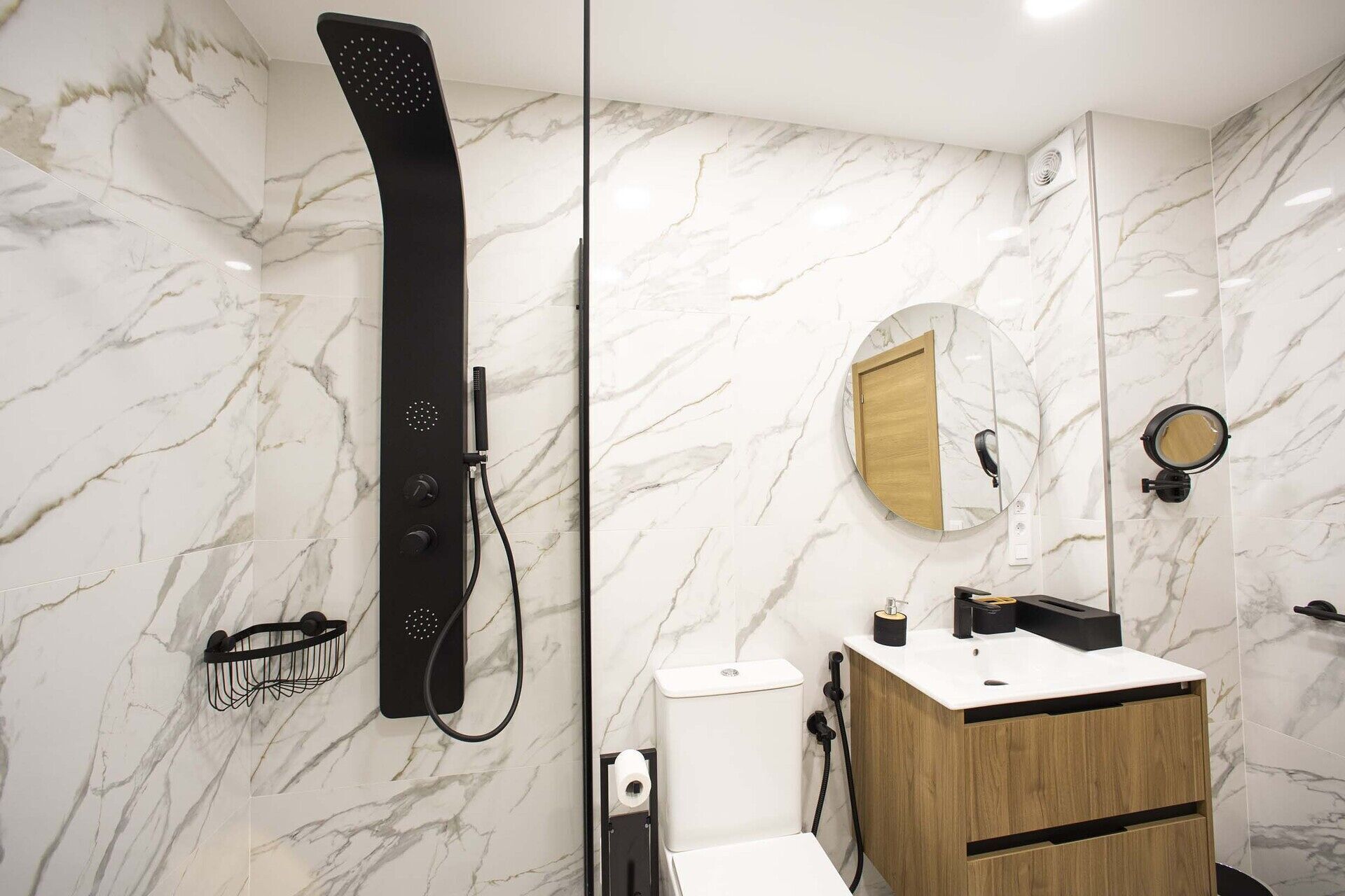 A modern bathroom with a marble wall, a black showerhead, a wooden vanity, and a round mirror.