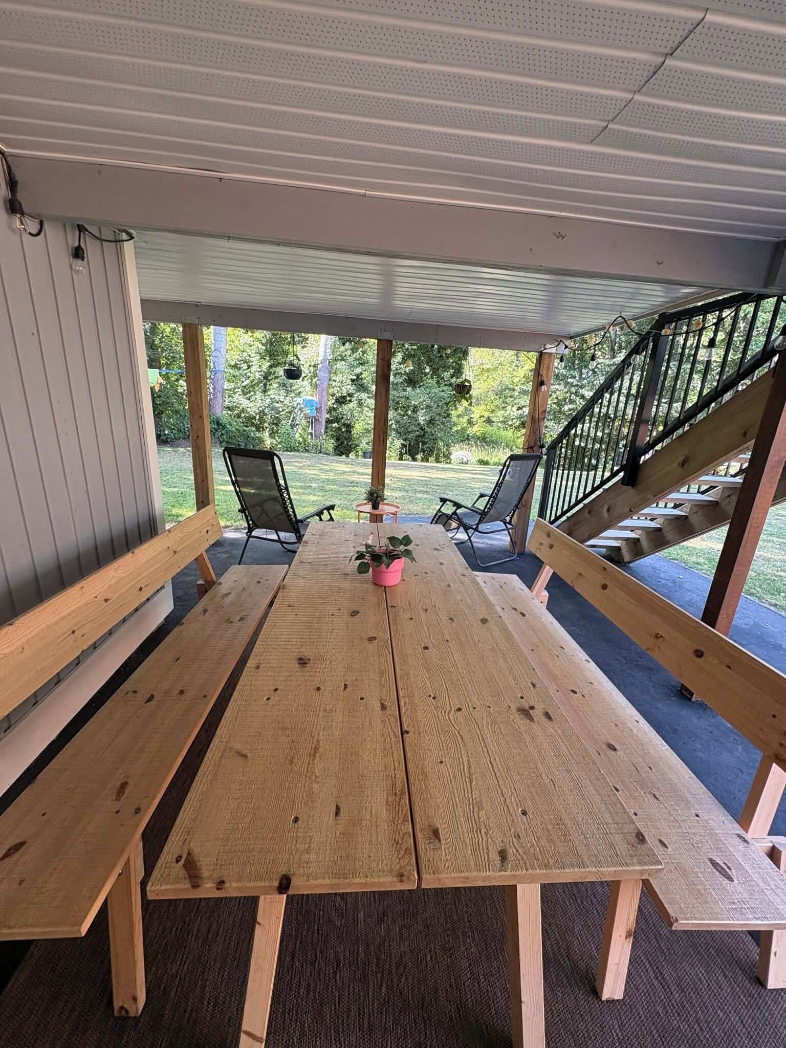 Un patio techado al aire libre con una mesa y bancos de madera, rodeado por una barandilla y una escalera.