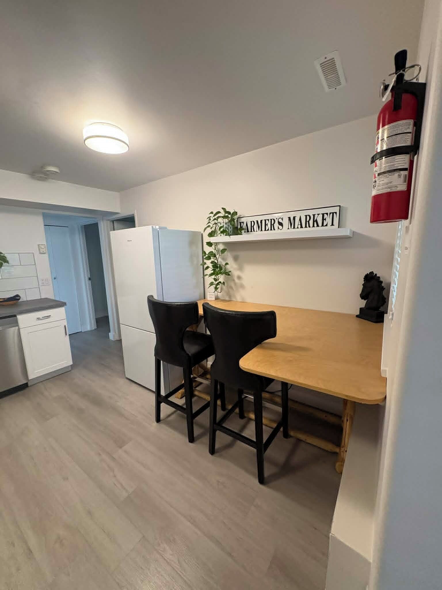 A small kitchen with a wooden table and two chairs, a sign reading 'FARMER'S MARKET', and a fire extinguisher mounted on the wall.