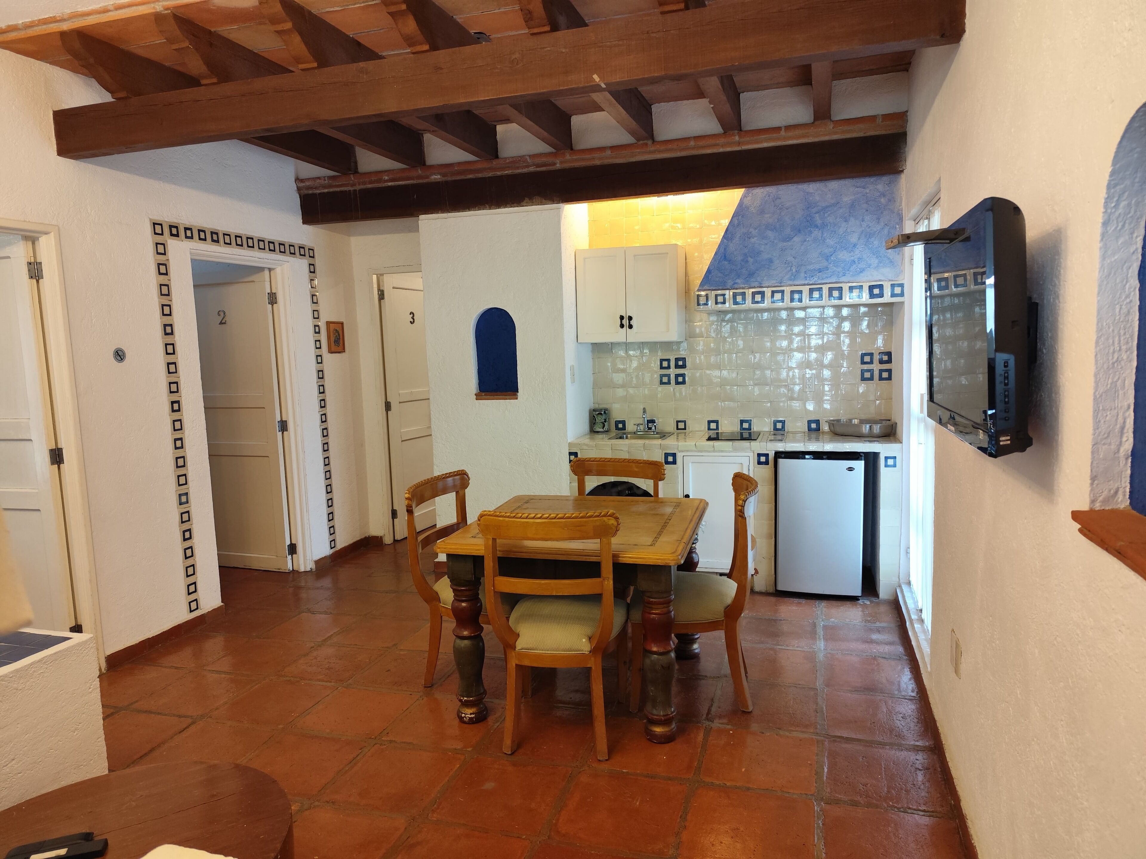 Zona pranzo con tavolo e sedie in legno, cucina con zoccolo in piastrelle e televisore a parete.