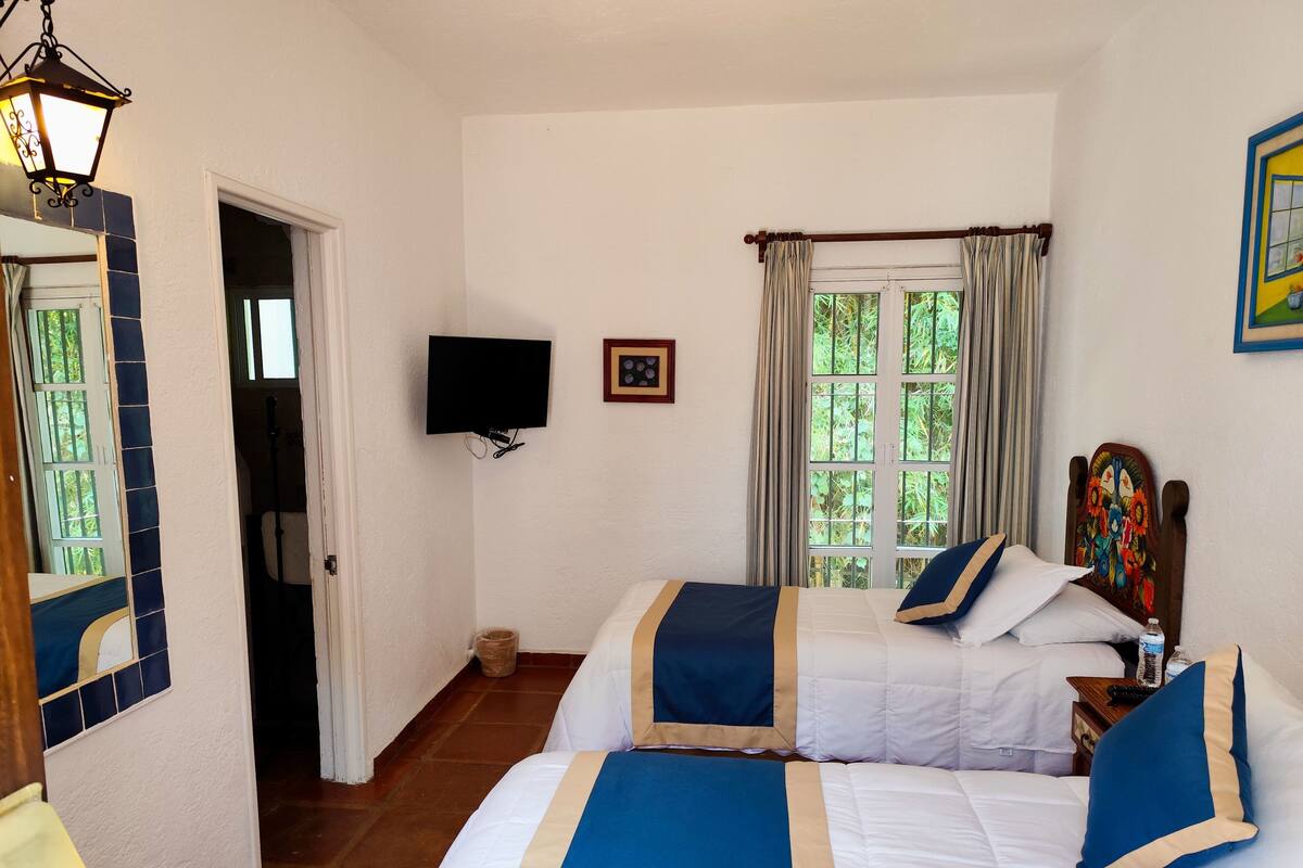 Quarto de hotel com duas camas, televisão, janela com cortinas e porta que dá para outro espaço.