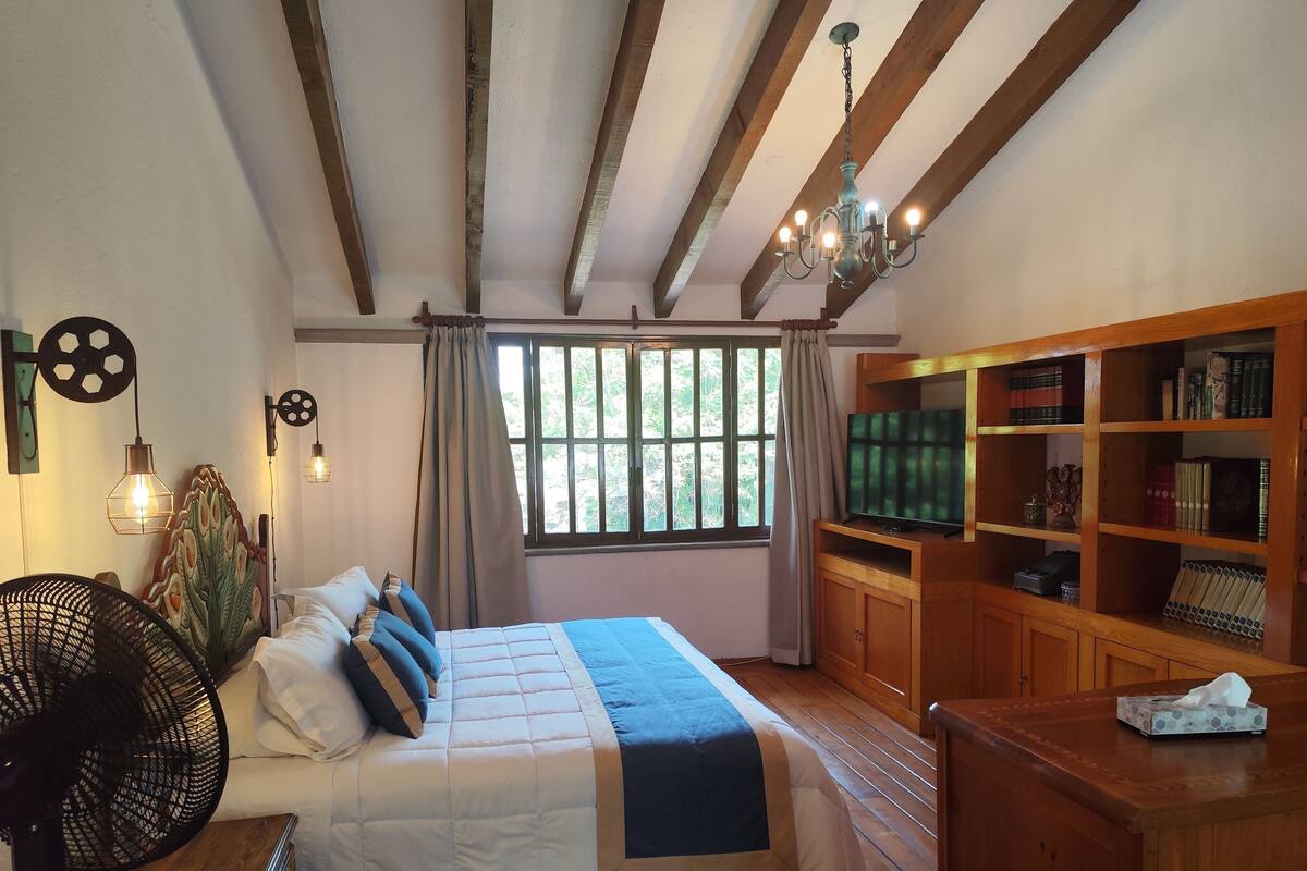 Quarto com uma cama grande, estantes de madeira, televisão e ventilador.