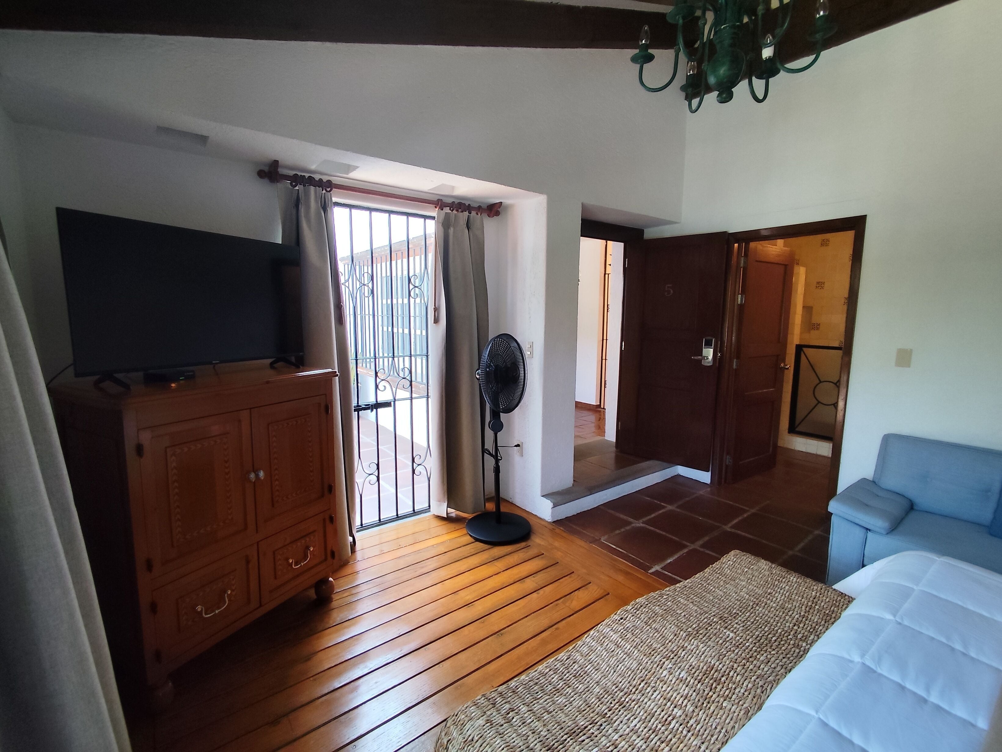 A room with wooden flooring, a television on a cabinet, a fan, a bed, and a door leading to another room.