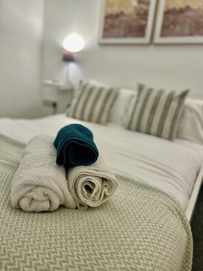 Un lit bien fait, avec des serviettes pliées et des coussins à rayures, une table de chevet avec une lampe, et deux tableaux encadrés accrochés au mur.