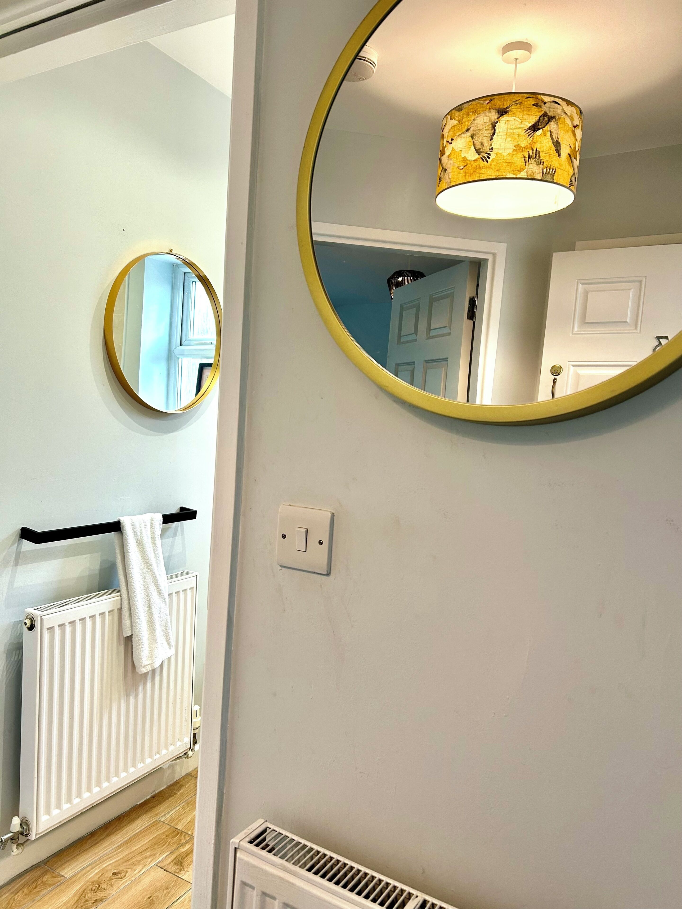 Ett badrum med ett vitt element, en handduk som hänger på ett svart handduksställ, en rund spegel med en gul ram och en lampa med en mönstrad lampskärm.