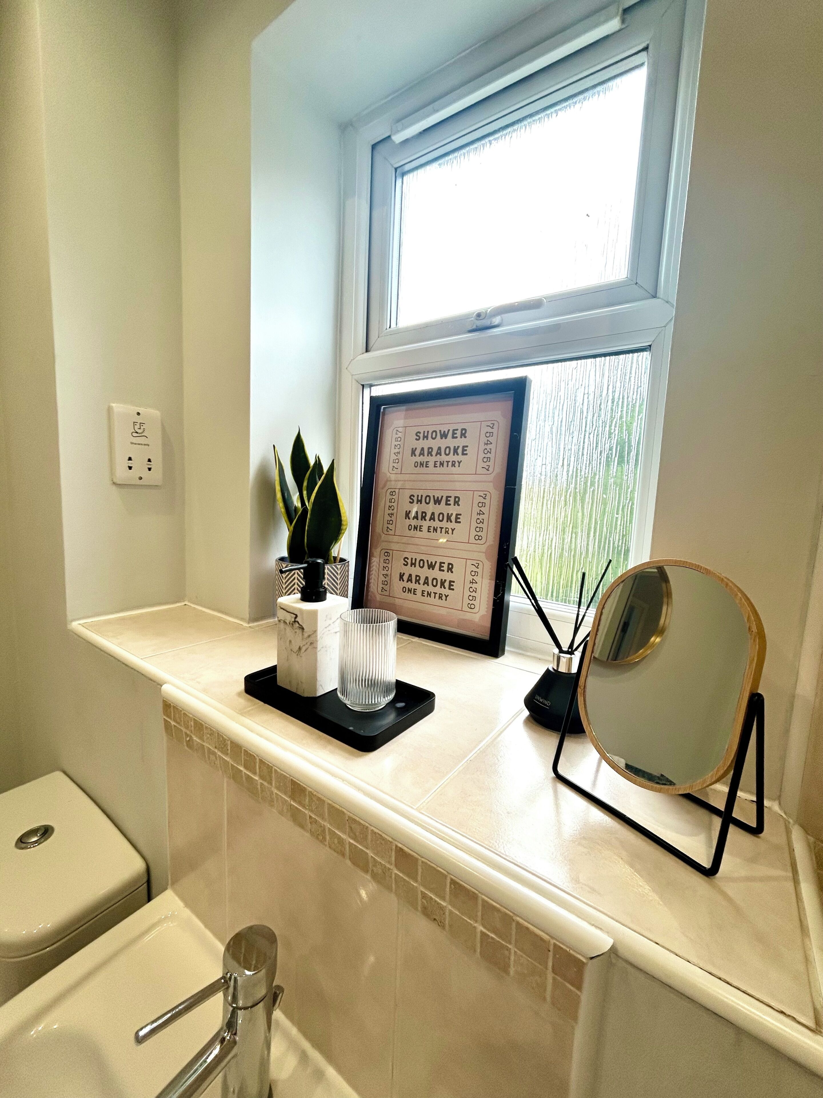 Une salle de bain avec une fenêtre, un panneau décoratif encadré, une plante, un verre et un miroir.