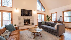 Ein großzügiges Wohnzimmer mit Kamin, einer breiten Couch, einem Couchtisch und großen Fenstern mit Blick ins Freie.