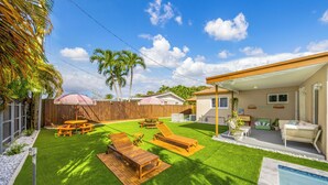 A backyard with artificial grass, wooden lounge chairs, a picnic table, and a pool.