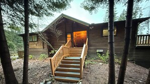 A wooden cabin with a staircase leading to a door, surrounded by trees and gravel.