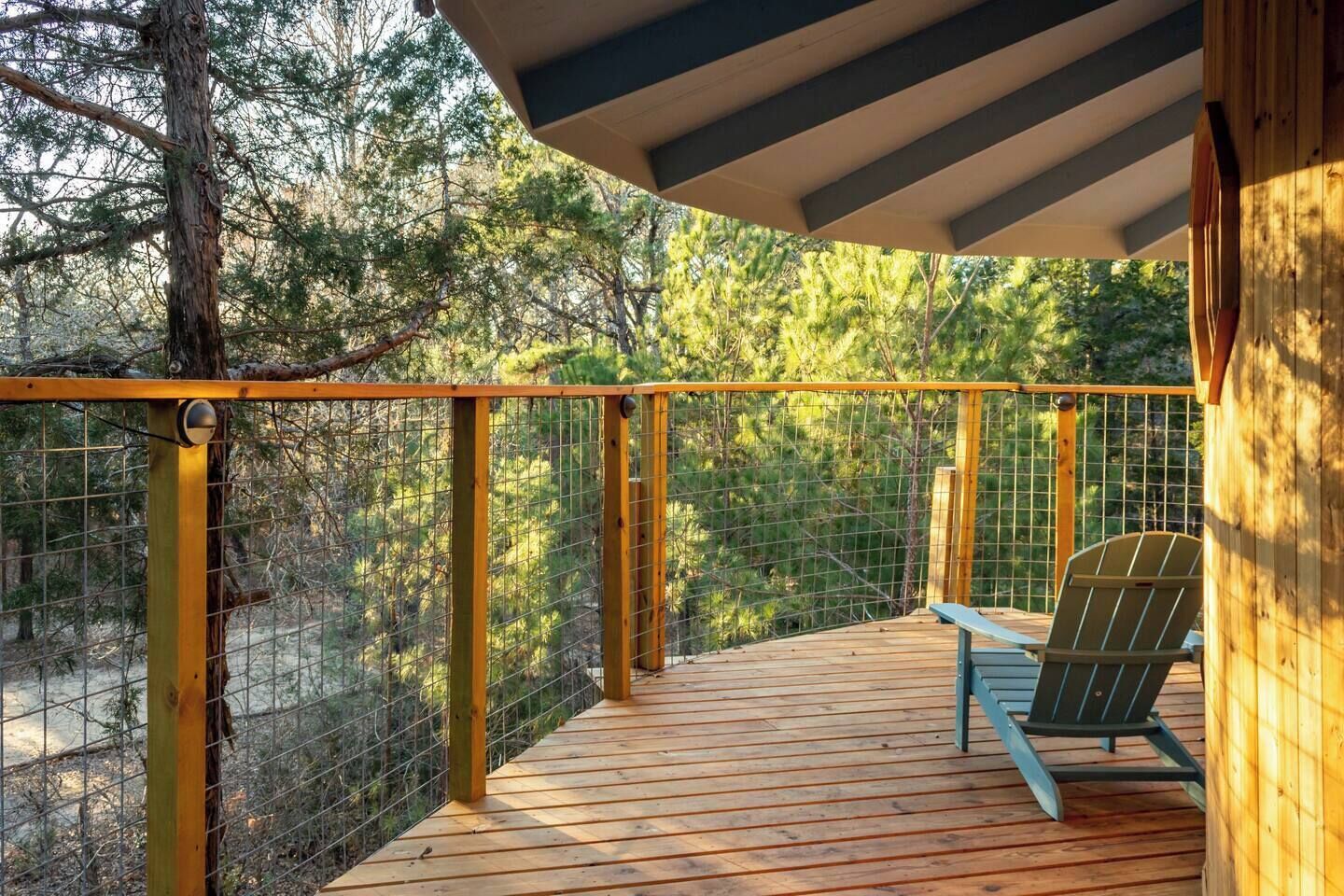 Una terraza de madera con barandilla metálica, una única silla Adirondack y vistas a un área boscosa.