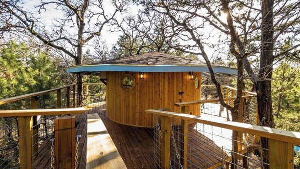 Une cabane en bois avec un toit bleu, entourée d’une terrasse en bois et de rampes en métal, située dans une zone boisée.