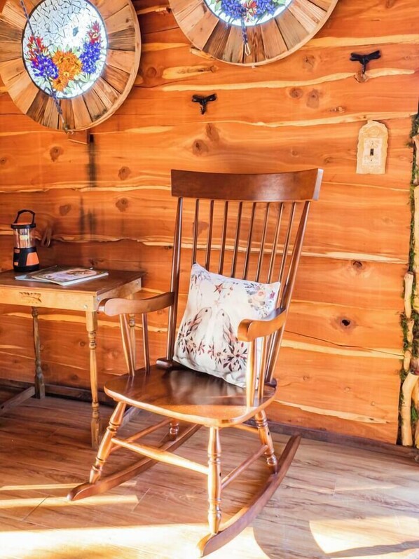 A wooden rocking chair with a floral cushion, a wooden table with a kettle, and a wall with two circular stained glass windows. - The Hobbit's Nest, A Magical Dog-friendly Treehouse in the Lost Pines Shire (Paige)