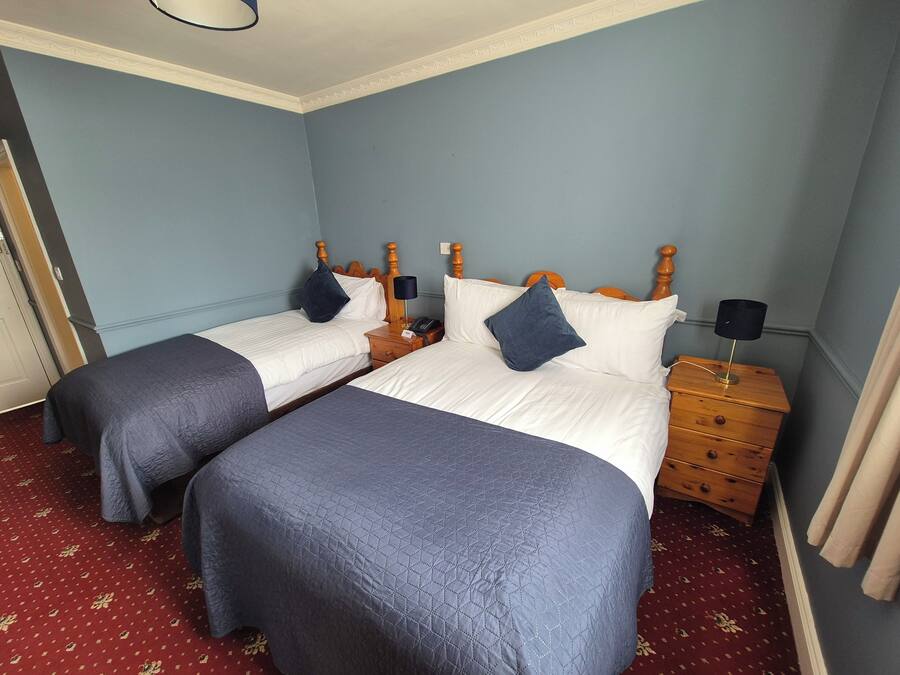 Une chambre d’hôtel avec deux lits, chacun doté d’une tête de lit en bois, de draps blancs et de coussins décoratifs bleus. On y trouve une table de chevet en bois avec une lampe et un téléphone. La chambre est dotée d’une moquette rouge et de murs bleu clair.