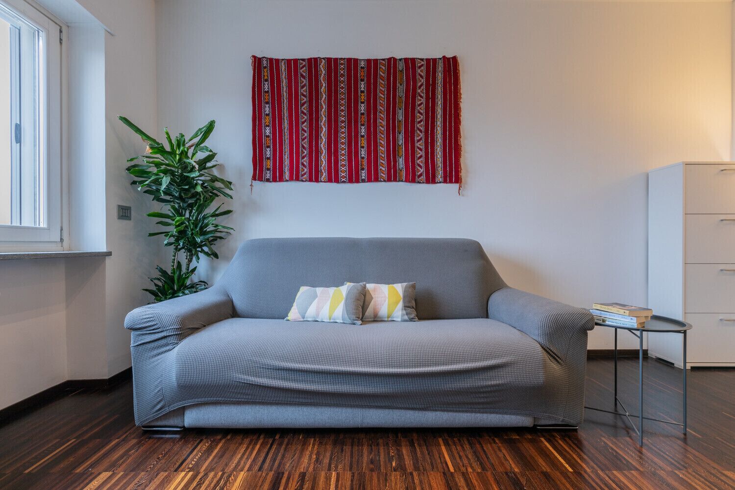 Un sofá gris con cojines estampados, un tapiz rojo y blanco, una planta en maceta y una pequeña mesa con libros.