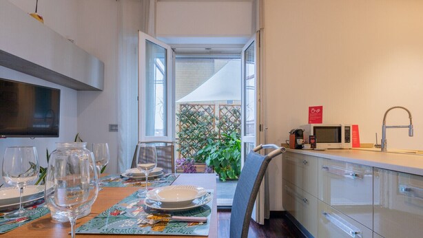 Uma sala de jantar com mesa posta para dois, uma cozinha com eletrodomésticos modernos e vista para um jardim através de uma porta aberta.