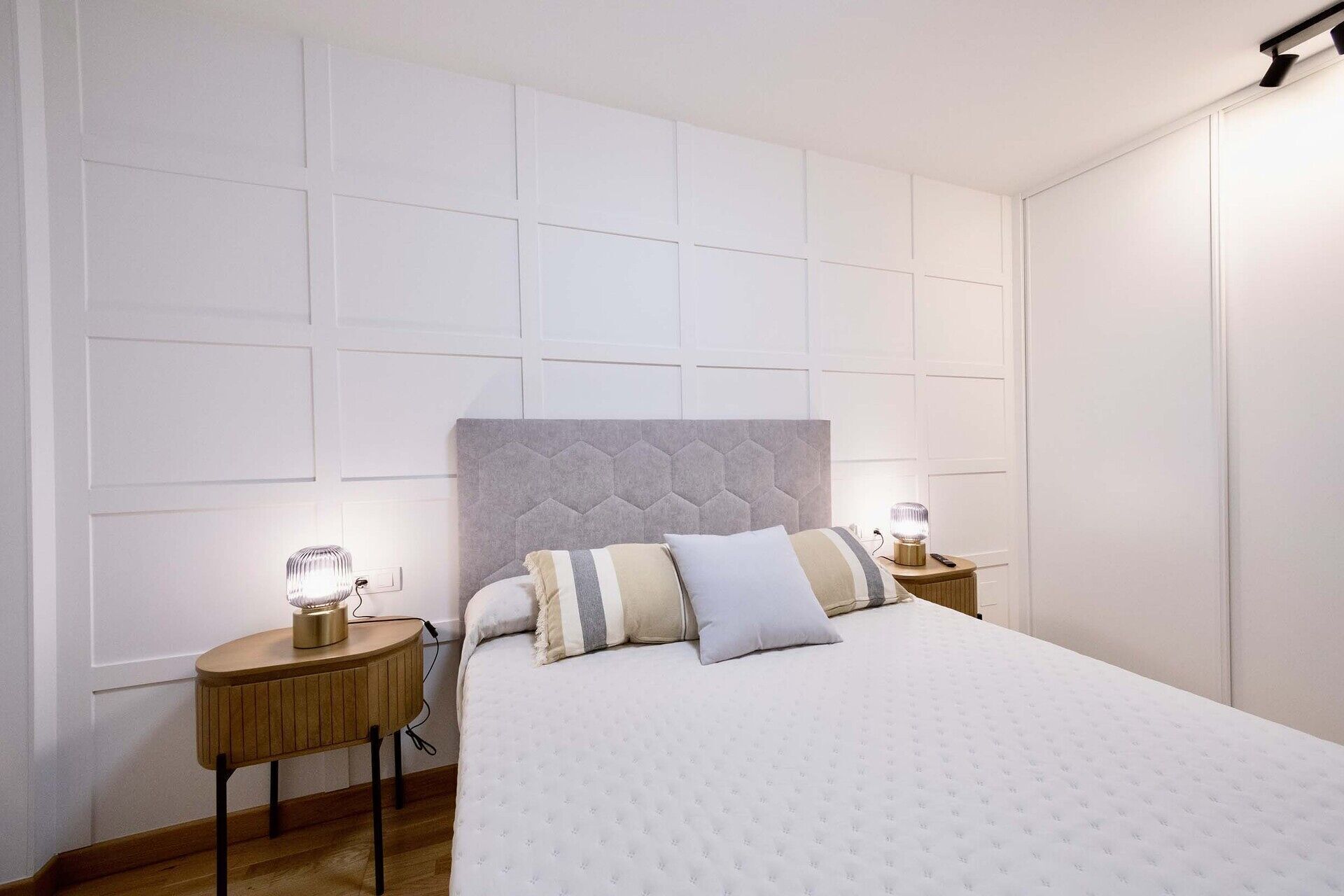 Een slaapkamer met een groot bed, twee nachtkastjes met lampen, een wit hoofdbord en een wandpaneel in wit design.