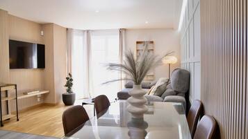 Een moderne woonkamer met een glazen eettafel, zachte zithoek, een flatscreen televisie en een groot raam.
