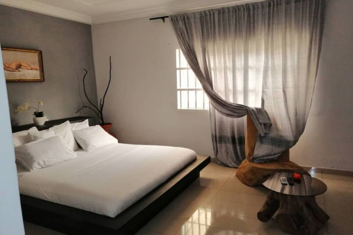 A modern bedroom with a large bed, a bedside table, a window with curtains, and a painting on the wall.