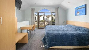 A bedroom with a bed, a desk, and a chair. There is a balcony with a view of houses.