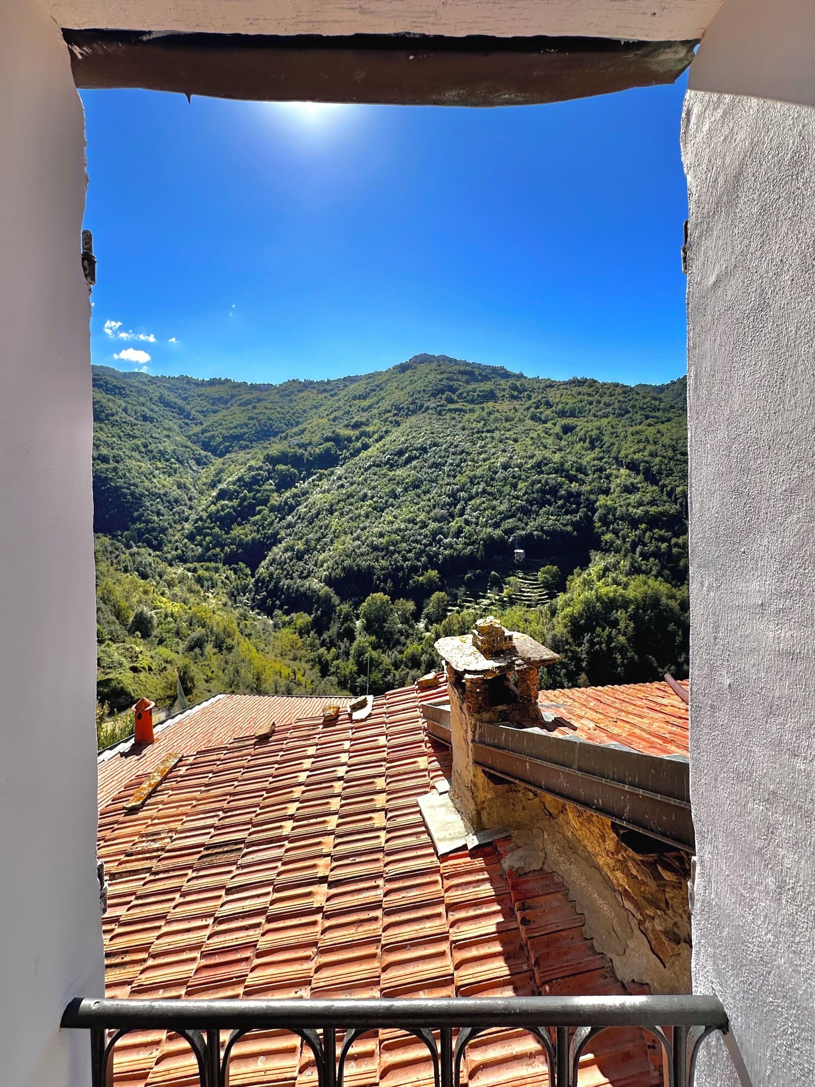 Vista da un balcone con piastrelle in terracotta, su una valle verdeggiante e una catena montuosa sotto un cielo azzurro limpido.