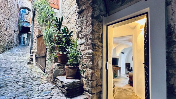 A narrow cobblestone alley with stone walls, potted plants, and a doorway leading to a room with a television and a chair. - Amazing place, solarium & Hamam (Apricale)