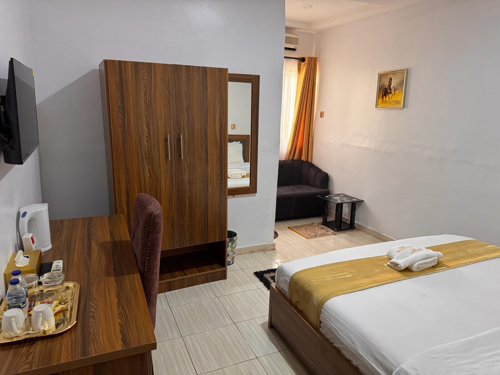 A hotel room with a wooden wardrobe, a bed with a white and gold bedspread, a small black table, a chair, and a television mounted on the wall.
