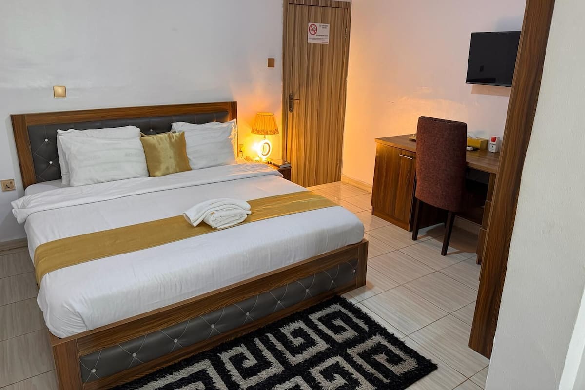 A neatly made bed with a headboard, a bedside table with a lamp, a wooden desk, and a chair.