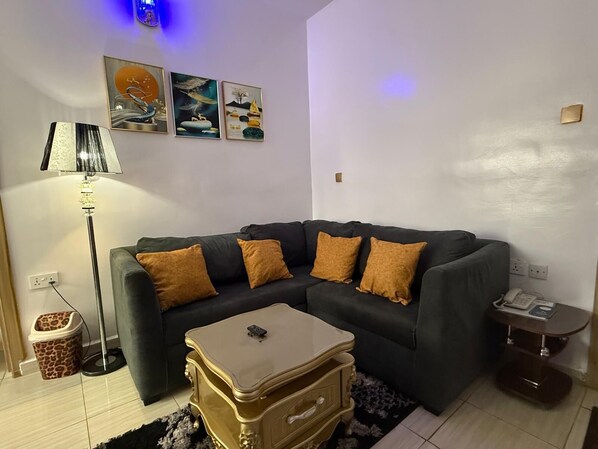 A living room with a dark green sofa, four mustard-colored cushions, a small beige side table with a remote control, a tall floor lamp, and a wall-mounted power outlet. - Akure Airport Hotel (Oba Ile)