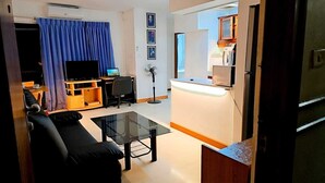 Un espacio habitable pequeño con un sofá, una mesa de centro de vidrio, un televisor, una computadora portátil y una zona de kitchenette.
