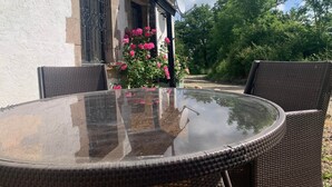 A round glass table with wicker chairs, a white building with barred windows, and pink flowers in the background.