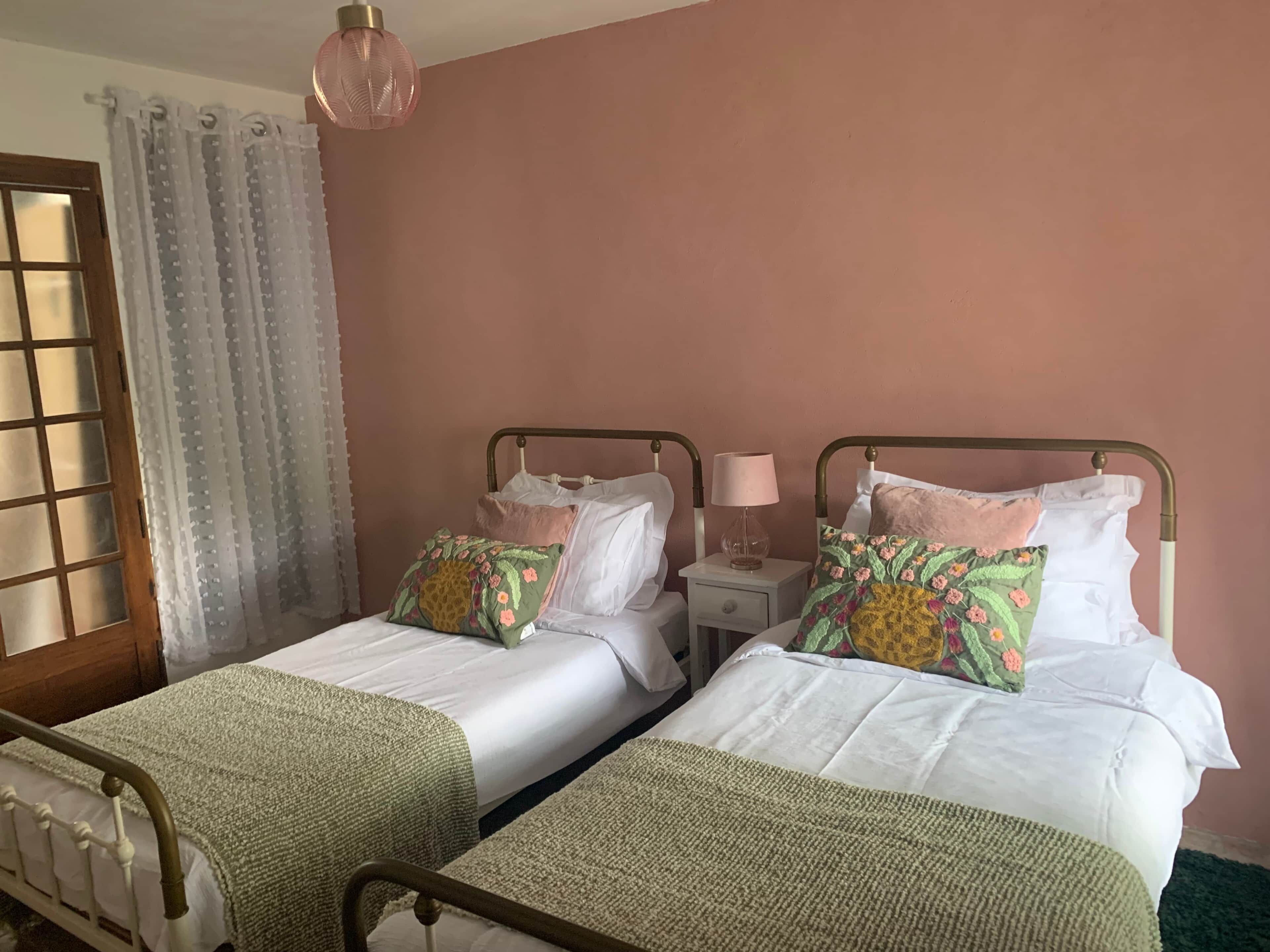Una habitación con dos camas, una mesita de noche con lámpara, una ventana con cortinas y una lámpara colgante.