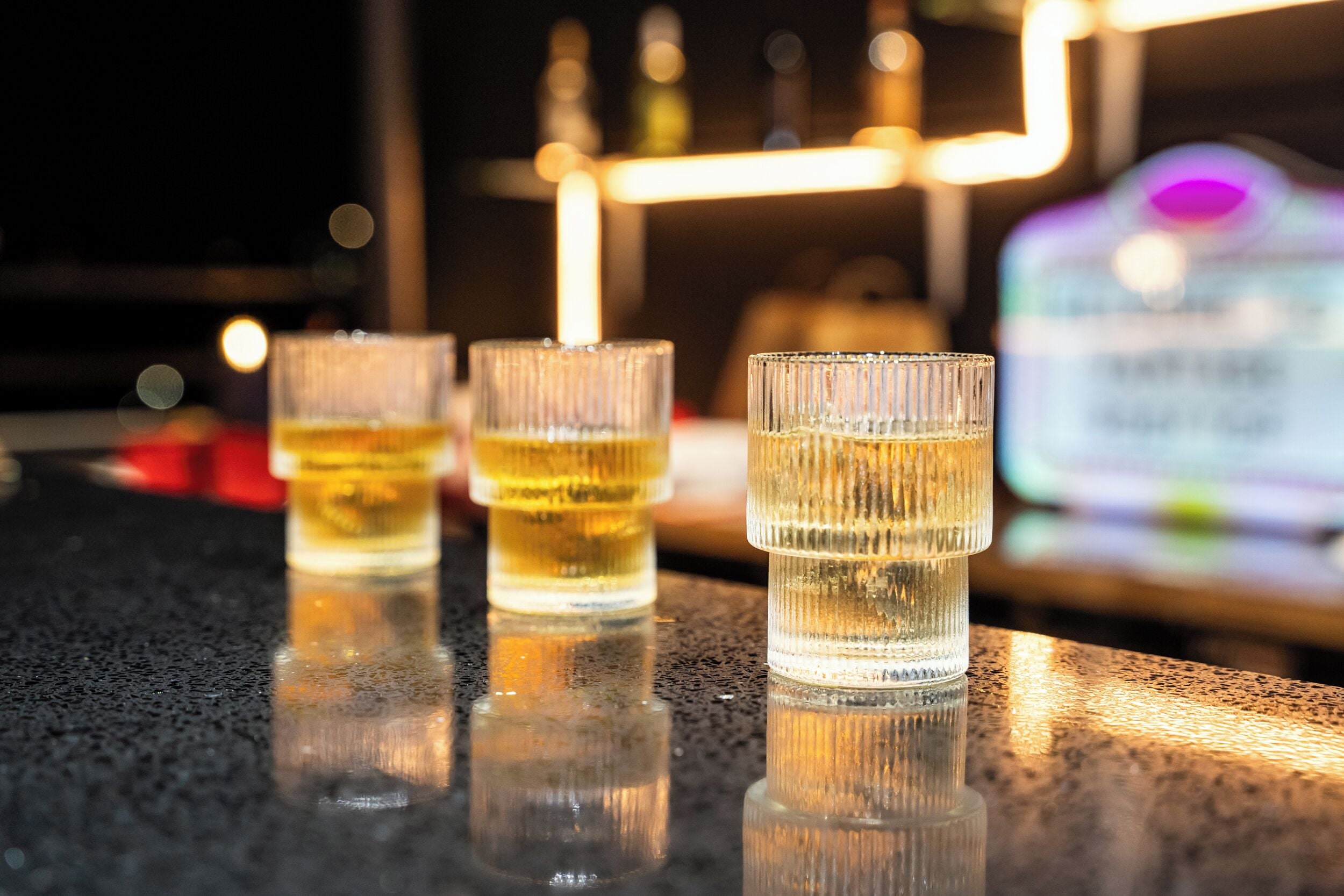 Three glasses of amber liquid on a reflective surface, likely a bar counter, with a blurred background featuring a neon sign and a bottle.