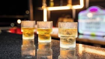 Three glasses of amber liquid on a reflective surface, likely a bar counter, with a blurred background featuring a neon sign and a bottle.