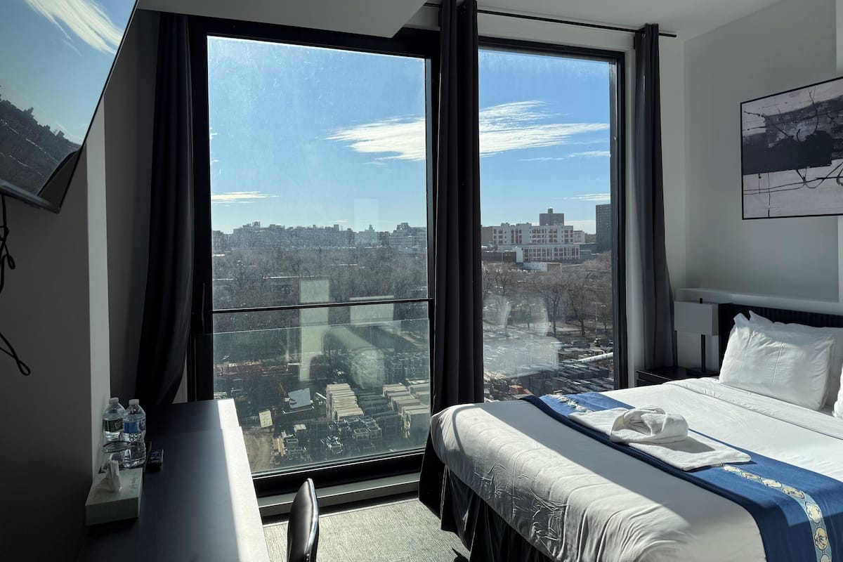 A hotel room with a large window offering a city view, a bed with white bedding, a bedside table with a bottle and a remote control, and a wall-mounted TV.