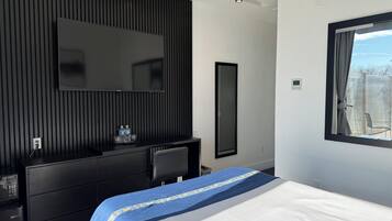 A modern hotel room with a large bed, a flat-screen TV mounted on a dark wall, a desk with a chair, and a window with a view of the outdoors.