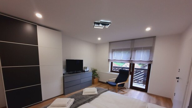 A modern bedroom with a large bed, a TV on a dresser, a chair, and a view of the outside through a window.