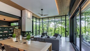 A modern interior with a dining area, sofa, and large windows offering a view of trees. - Modern new 4 bed 3 bath cottage on Bruce Lake, SE Exposure, 370ft frontage (Port Carling)