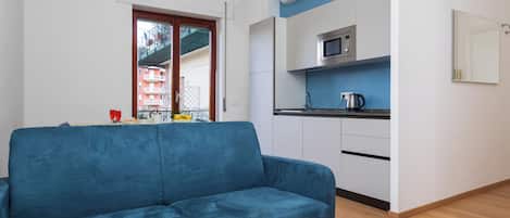 A modern living room with a blue sofa, a kitchen area with a microwave, and a window with a view of buildings.