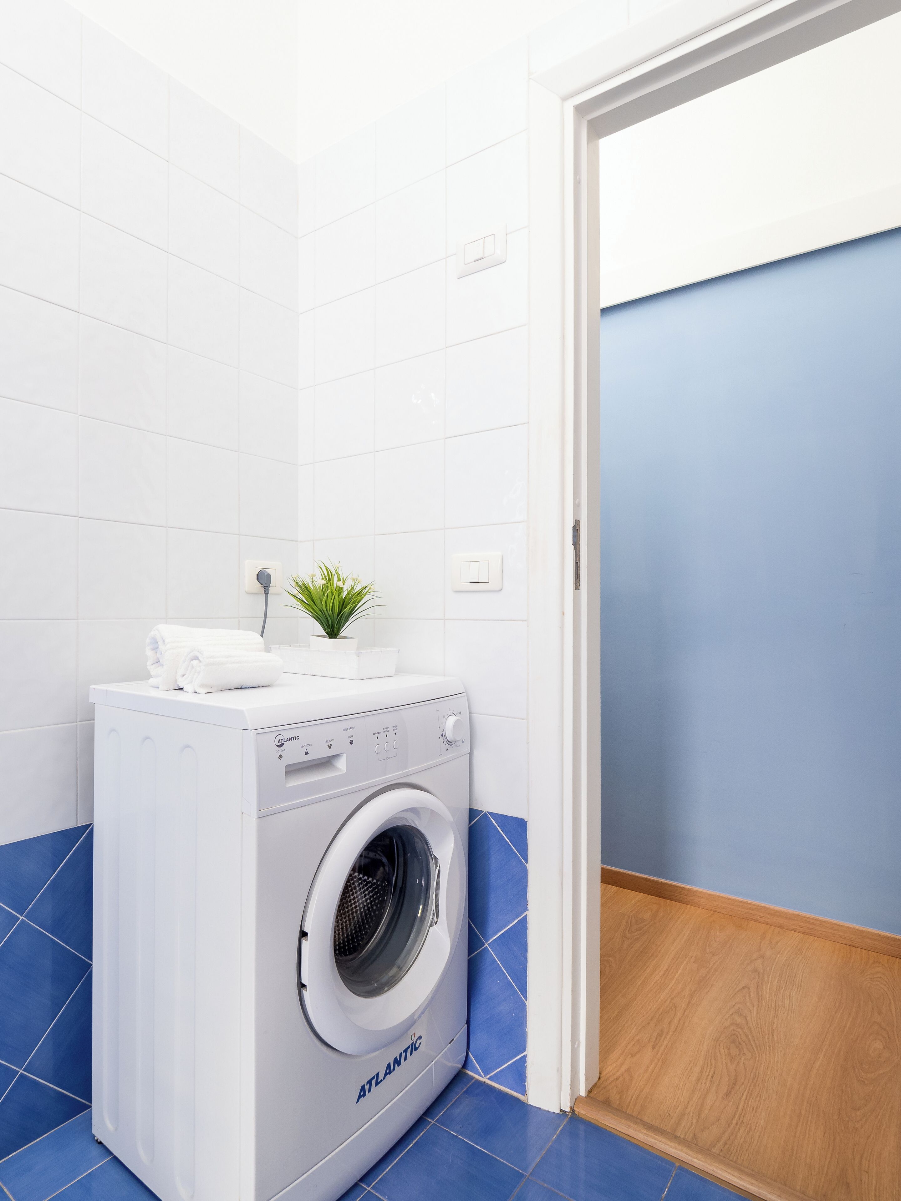 A compact, white washing machine with a small plant on top, set against a blue-tiled floor and white walls.