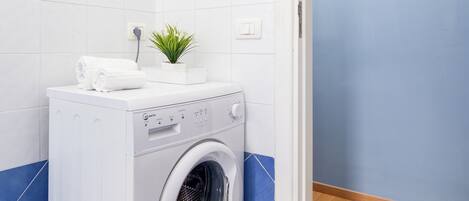 A compact, white washing machine with a small plant on top, set against a blue-tiled floor and white walls.