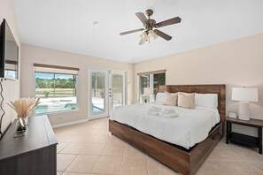 Ein Schlafzimmer mit einem großen Bett, einem Deckenventilator, einem Nachttisch, einer Lampe und Blick auf einen Swimmingpool durch eine Glastür.