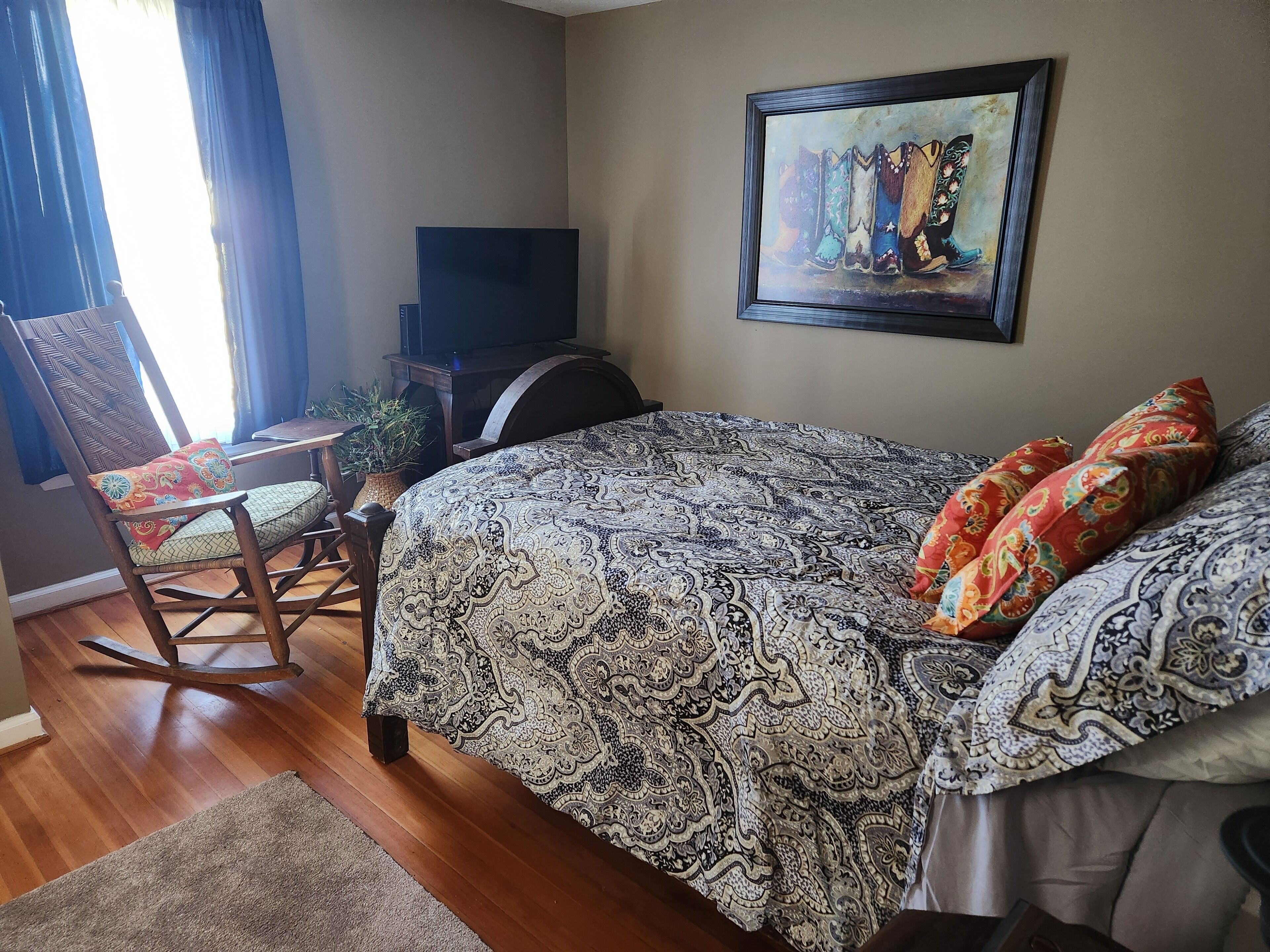 A bedroom with a patterned bedspread, a rocking chair, a television, and a framed artwork on the wall.