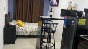 A compact room with a bed, a small desk with a chair, a refrigerator, and a television mounted on the wall.