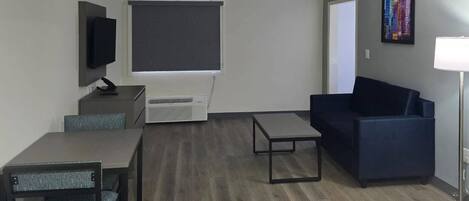 A modern living room with a dark blue sofa, a small table, a TV on a stand, and a large window with blinds.