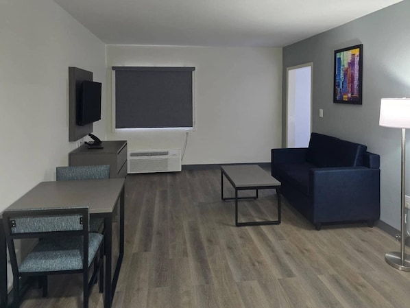A modern living room with a dark blue sofa, a small table, a TV on a stand, and a large window with blinds. - MainStay Suites Prince George - Fort Lee (Prince George)