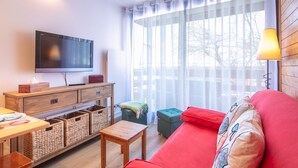 A modern living room with a flat-screen TV, wooden furniture, a red sofa, and a large window with sheer curtains. - Le Bleuet - Vue montagne, pistes accessibles à pied (Chamrousse)