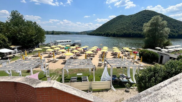Ein See mit Strand, Liegestühlen und Sonnenschirmen, ein Boot liegt im Wasser und im Hintergrund ist ein Berg zu sehen.